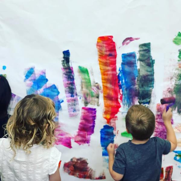 Children painting colorful strokes on a large white canvas.