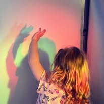 Child with multicolored shadow on wall, reaching up.