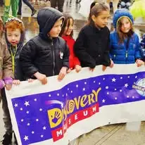 Children in jackets holding a Discovery Museum banner on a rainy day.