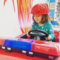 Child in a red fire hat playing in a toy fire truck, holding the steering wheel.
