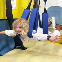 Child smiling, suspended in blue harness with yellow wall background, arms outstretched like flying.