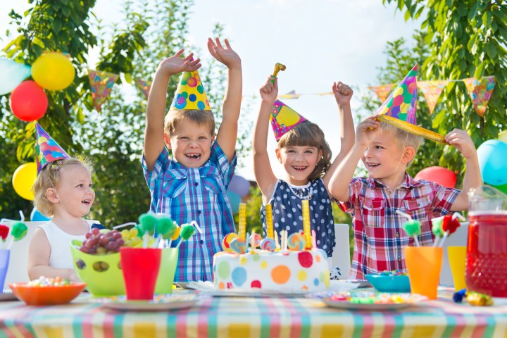 Four children in party hats celebrating outdoors with a cake and decorations.