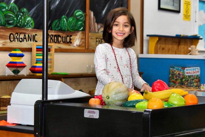 Smiling child at a play grocery store with toy produce and cash register.