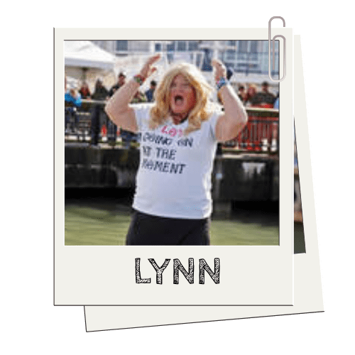 Person with long hair in a T-shirt cheering outdoors with arms raised.
