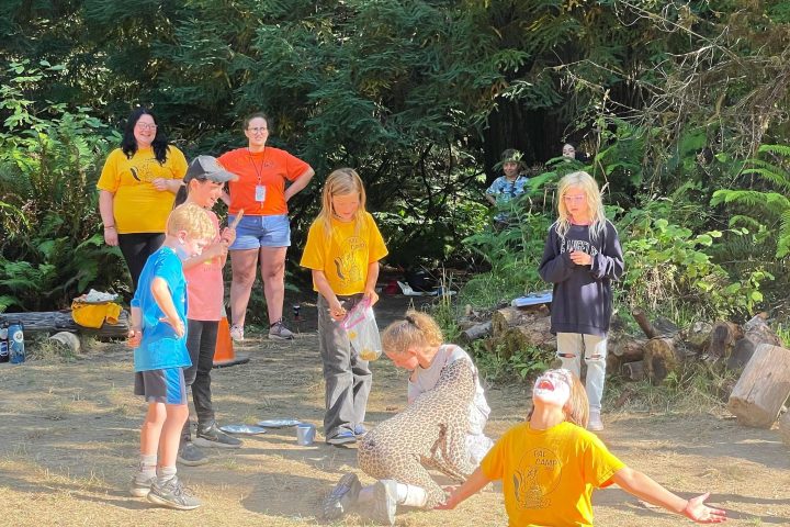 Children play in a sunny forest setting, some kneeling and one with white face paint.