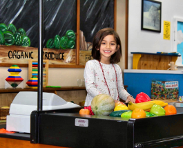 Girl playing at a toy cash register with plastic fruits and vegetables in a pretend grocery store.