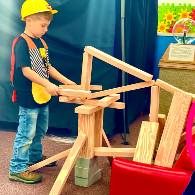 Child in a hard hat builds with wooden blocks in a play area.