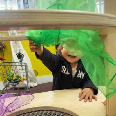 Child playing peek-a-boo with a green scarf in a play area with a small shopping cart in the background.
