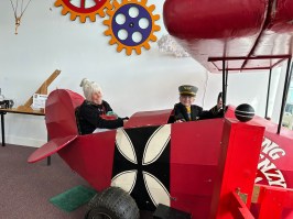 Child and woman sitting in a red toy airplane with large colorful gears on the wall behind them.