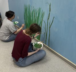 Two people seated on the floor, painting grass on a blue wall in an art space.