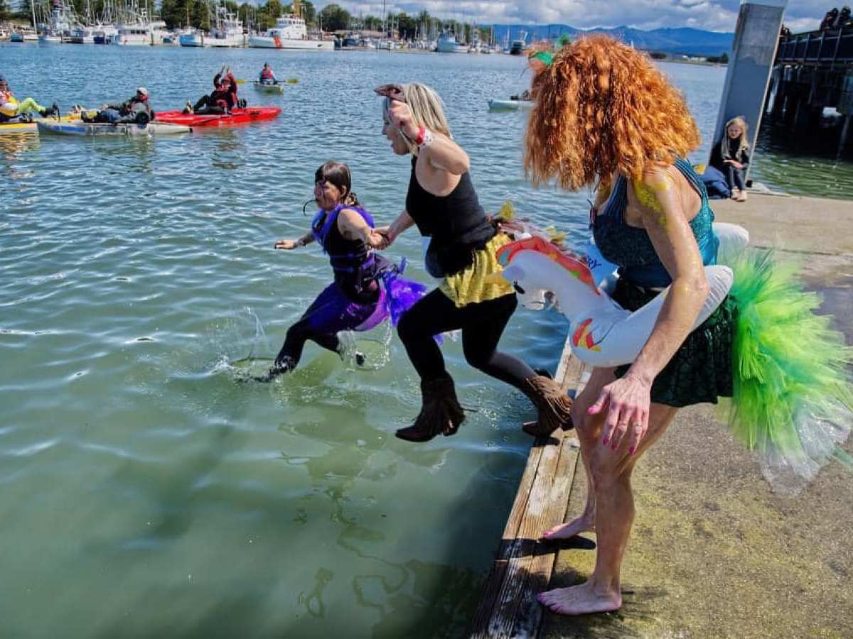 Three people in colorful costumes jumping into water near kayaks and boats.