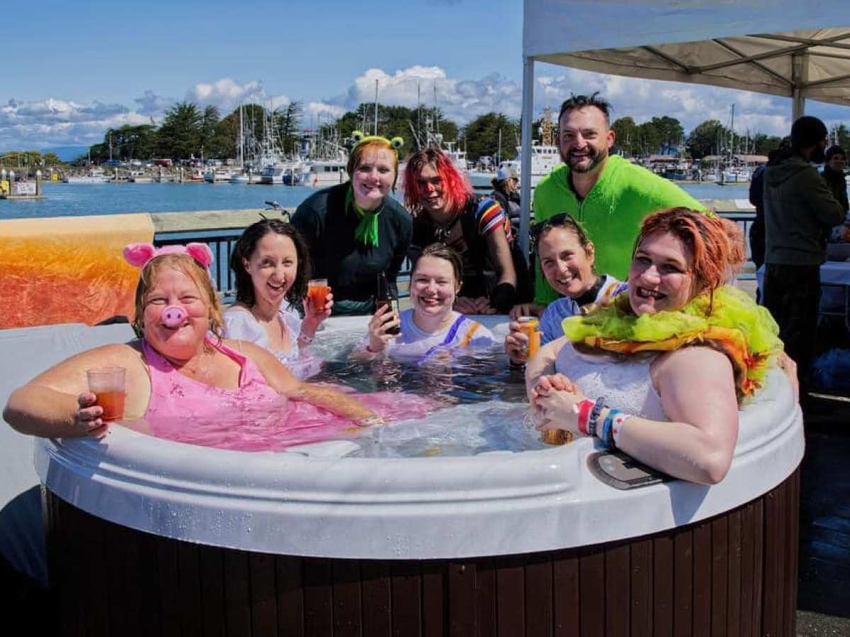 Seven people in costumes enjoying drinks in a hot tub by the waterfront.