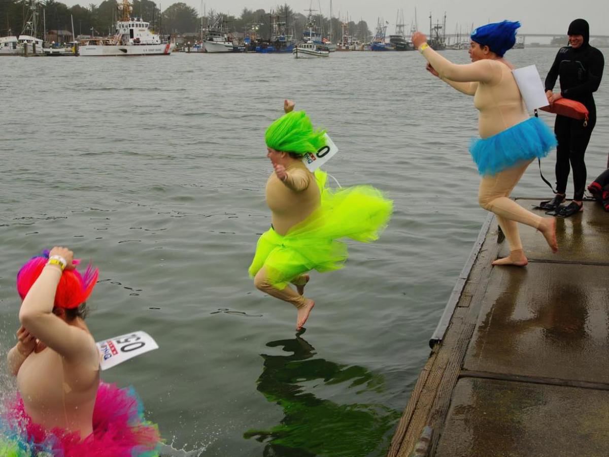 Three people in colorful wigs and tutus joyfully jump into a harbor, with boats in the background.