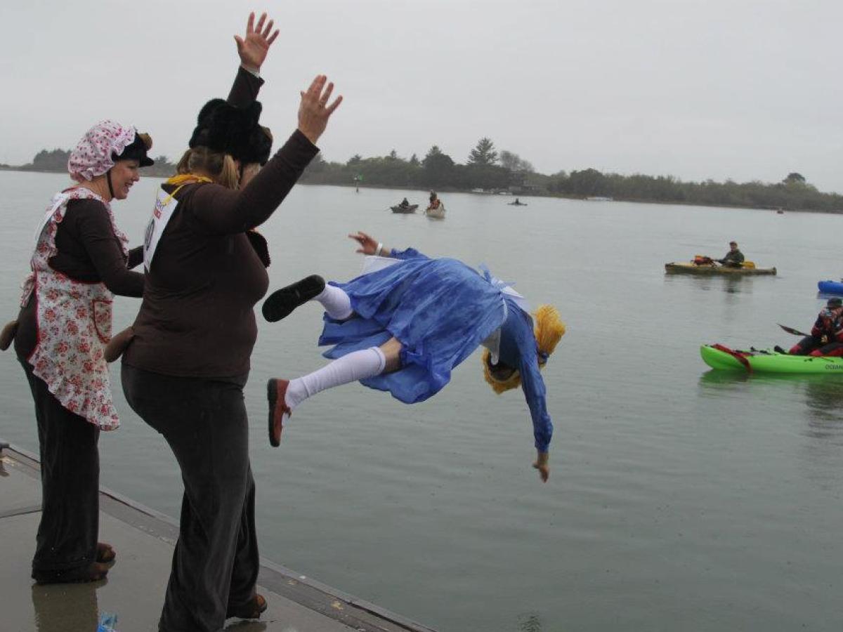 Person in blue dress being playfully thrown into water with onlookers watching.