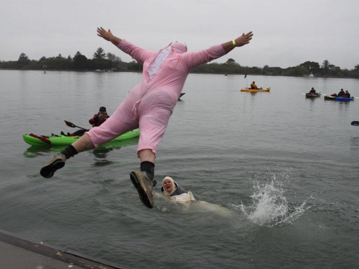 Person in pink costume jumps into lake, surrounded by kayakers.