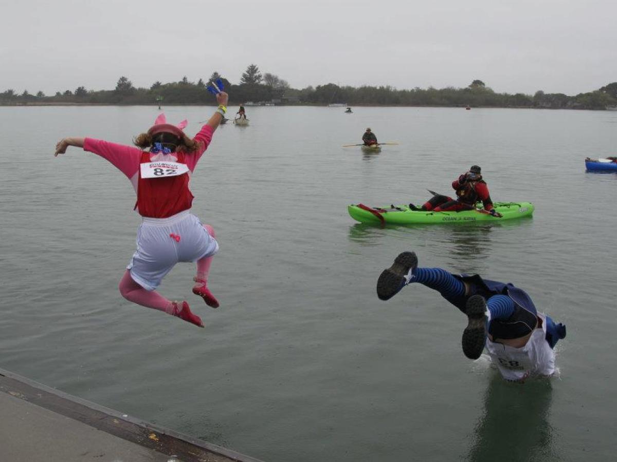 Two people in costumes jump into a lake while others kayak nearby.