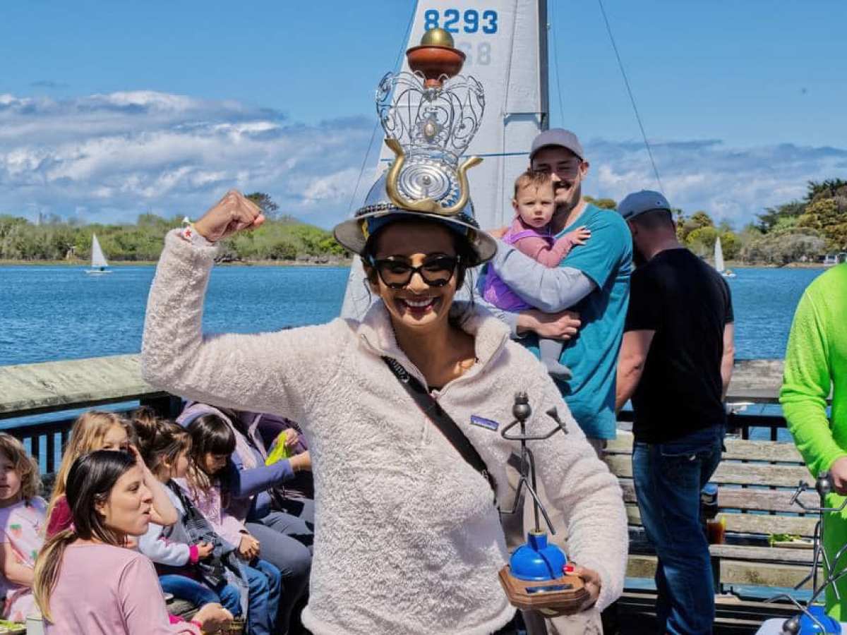 Person smiling and flexing with unique hat, holding a trophy by a lake, surrounded by people.
