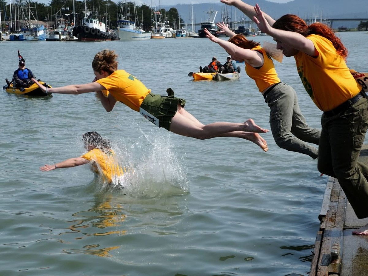 Three people in yellow shirts jump off a pier into water with kayaks in the background.