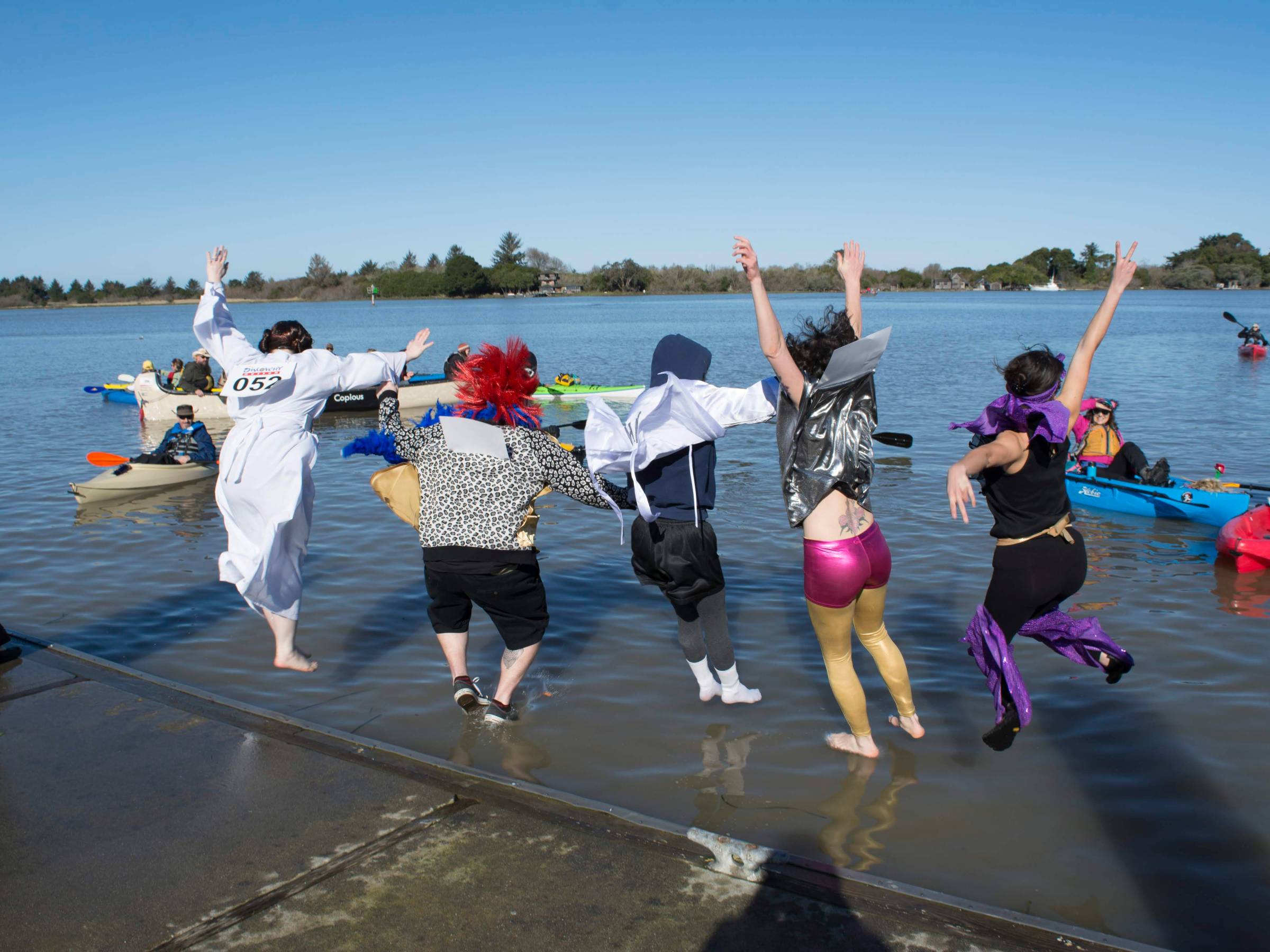 Costumed people jumping into a lake with kayakers in the background on a sunny day.