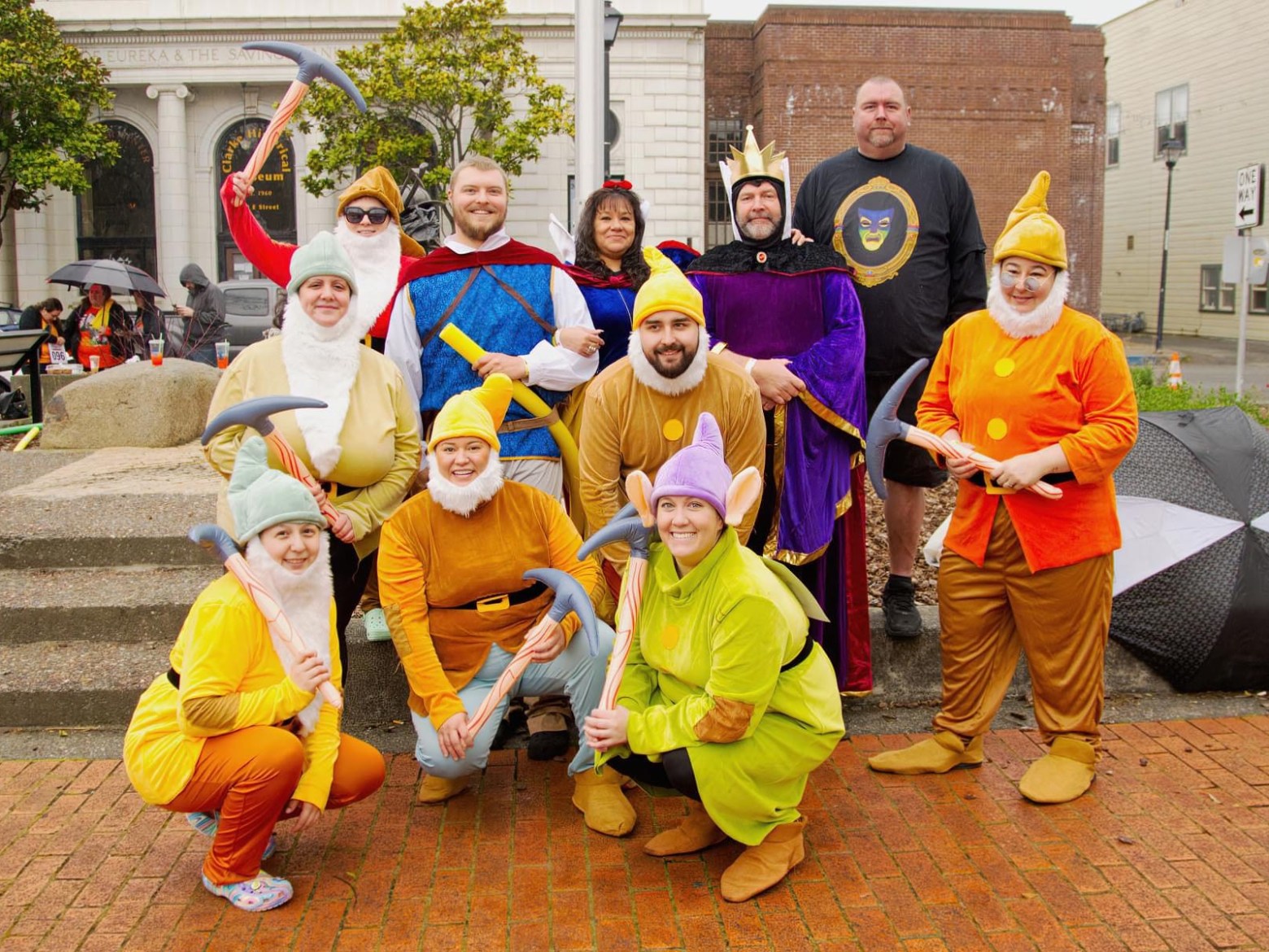 Group in colorful costumes resembling fairy tale characters, posing outdoors with props like pickaxes.
