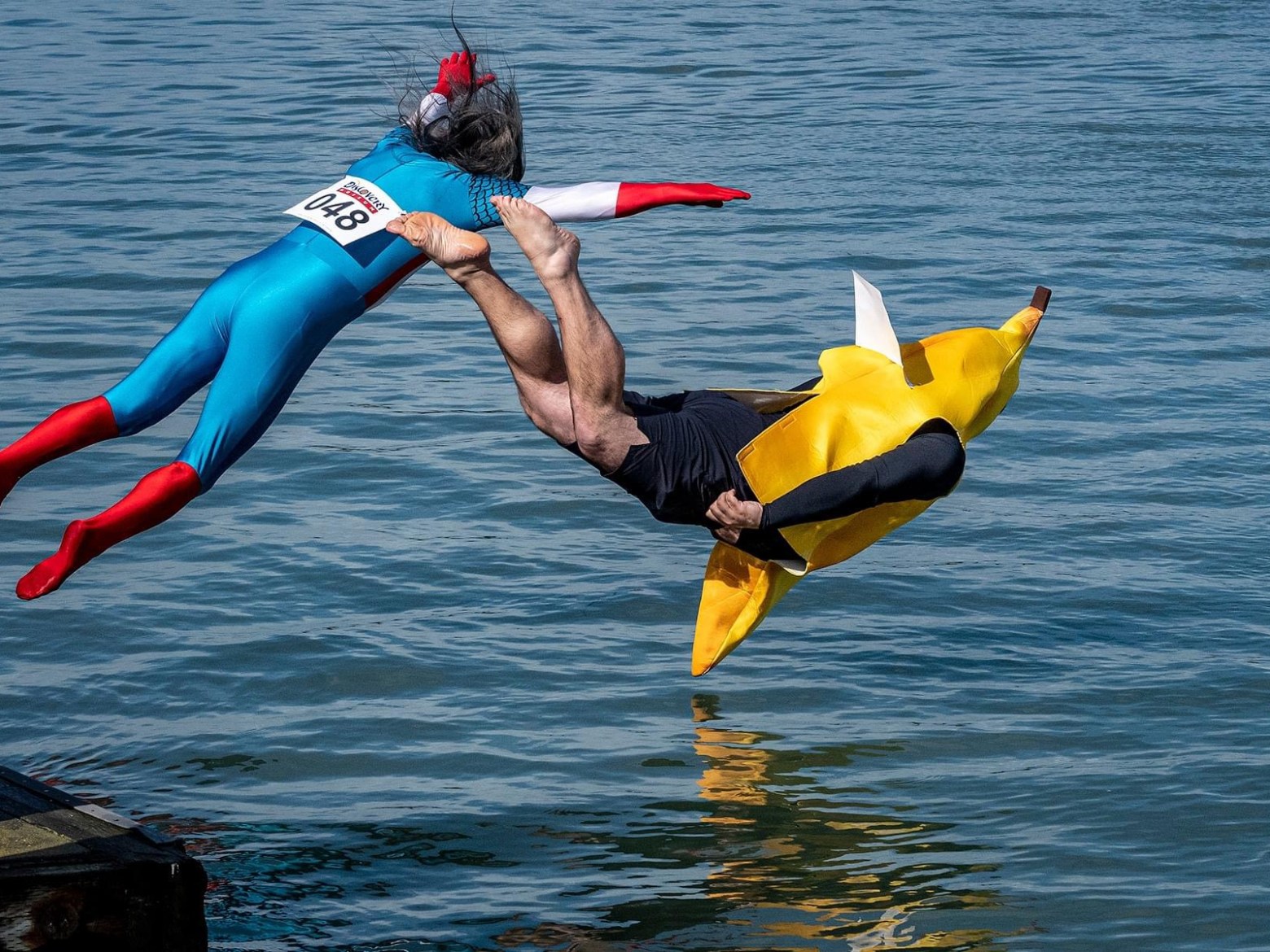 Two people in costumes jump into water; one in superhero suit, the other in a banana outfit.