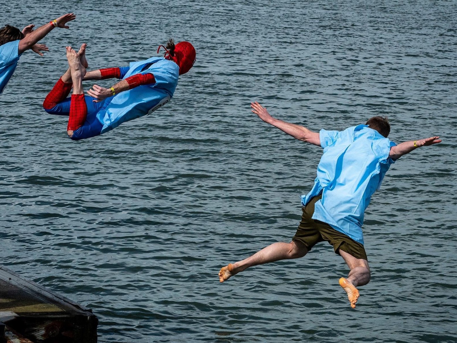 Three people in blue shirts jump into a body of water, one in a red superhero costume.
