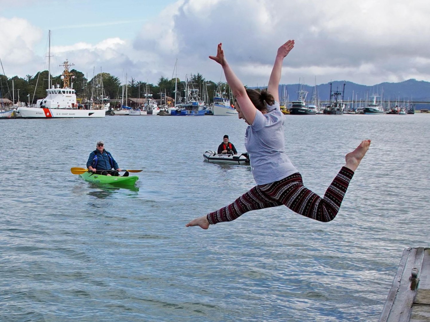Person jumps into water near kayaker and boat in harbor.