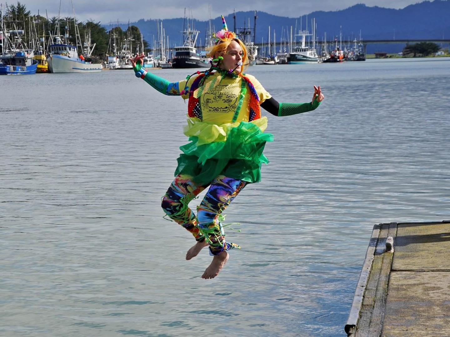 Person in colorful costume jumps off dock into water with boats in the background.