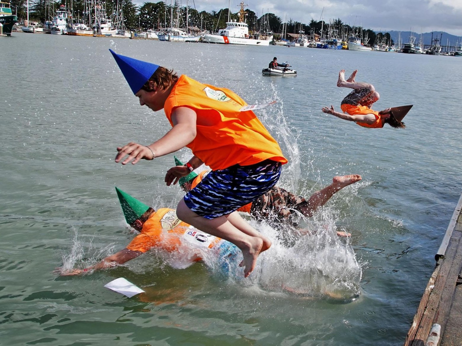 People in orange shirts with hats jump into a harbor, creating splashes.