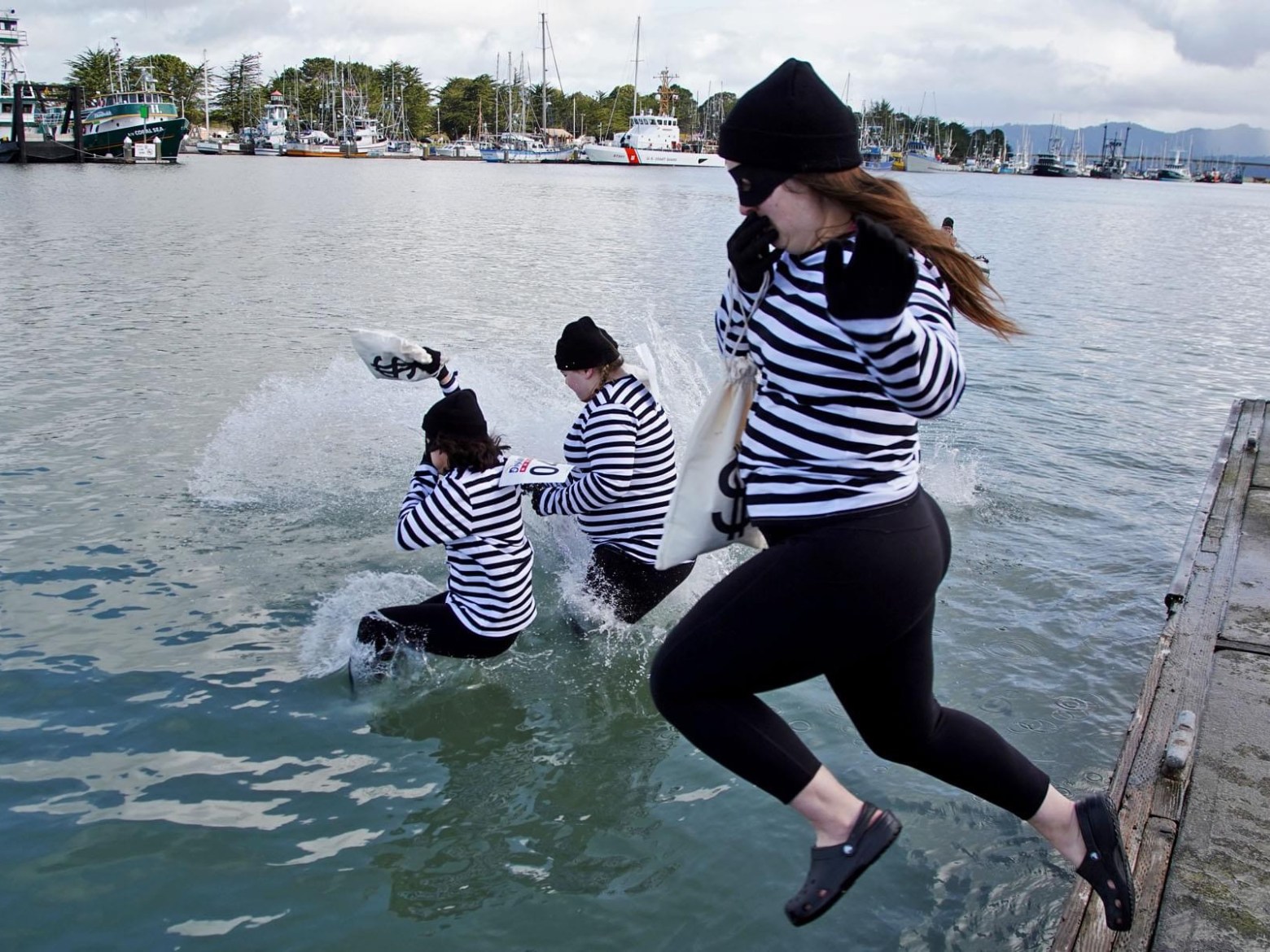 Three people in striped shirts jump into water from a dock, dressed as burglars with fake money bags.