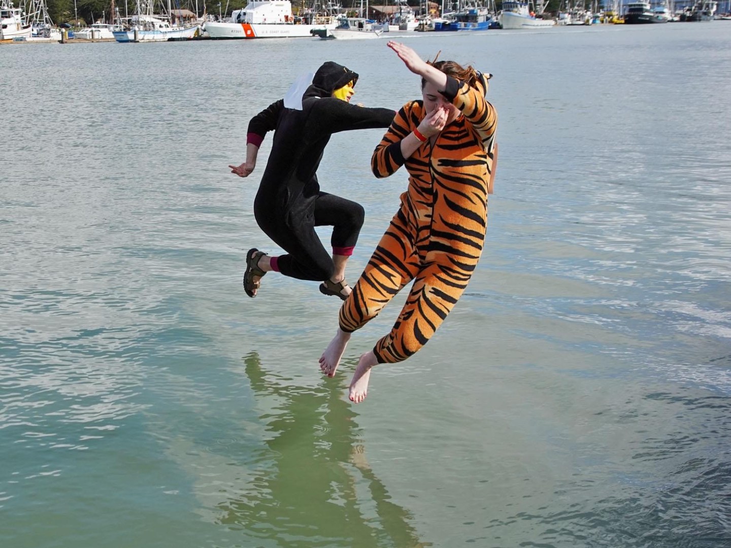 Two people in animal onesies jumping into water with boats in the background.