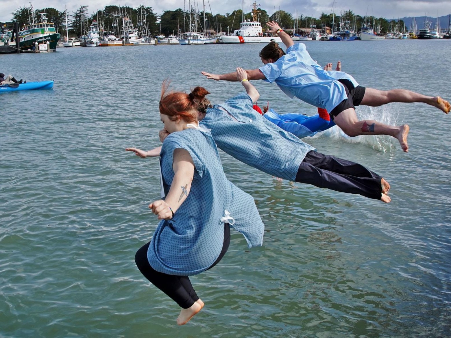 People in hospital gowns jumping into a harbor with boats in the background.