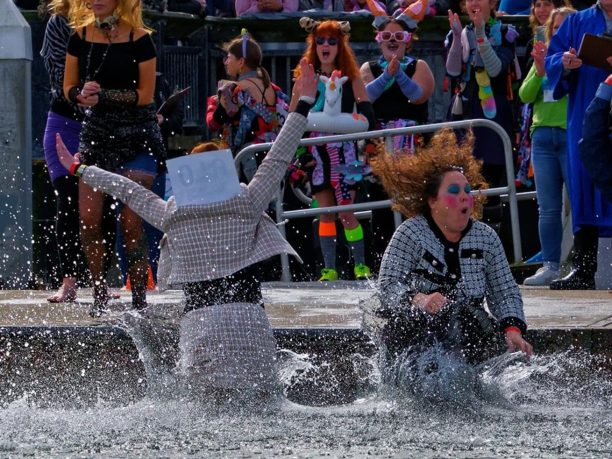 Two people in costumes splash into water while onlookers cheer and clap.