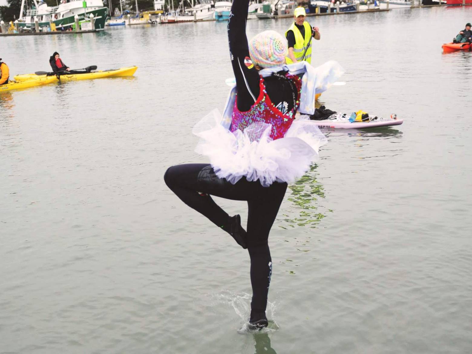 Person in colorful outfit and tutu performs on water with kayakers and paddleboarder in background.