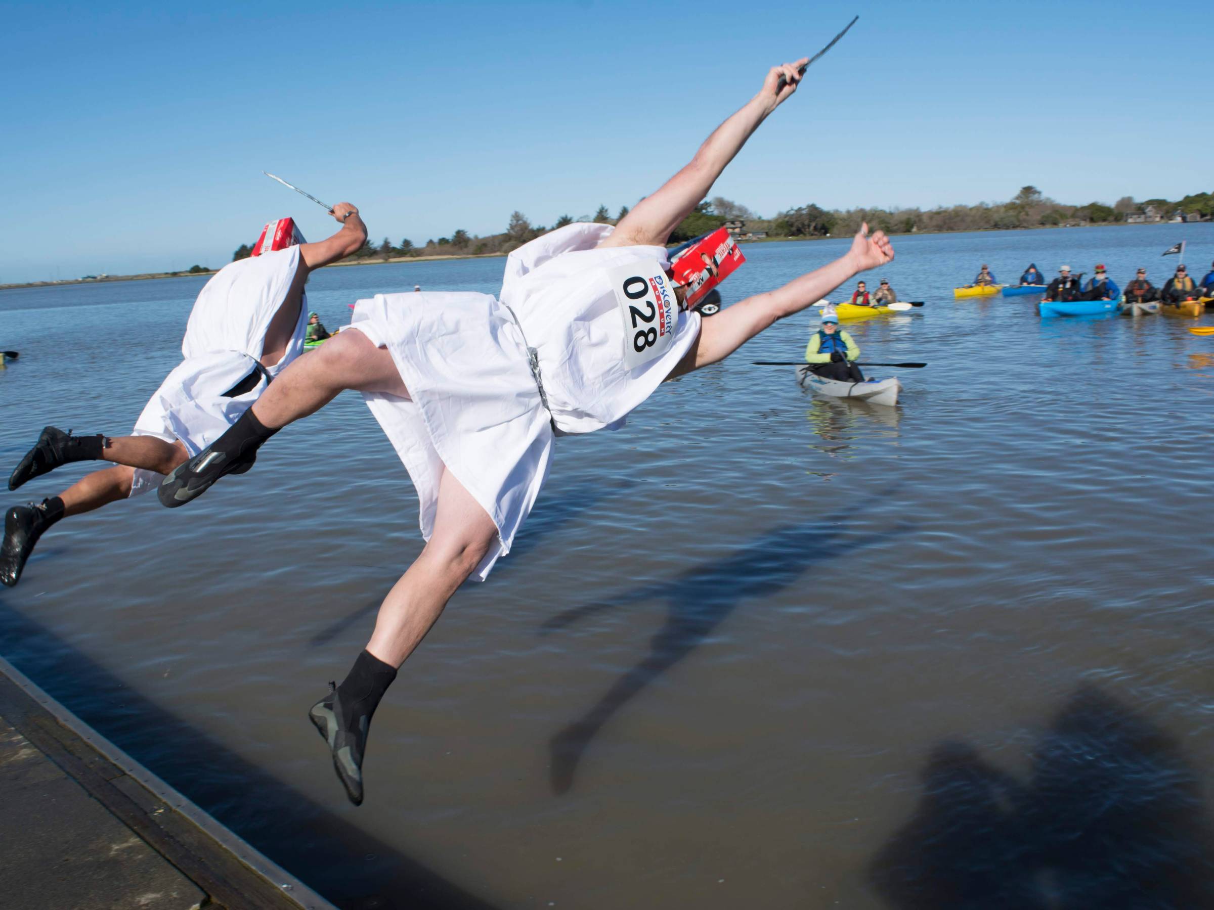 Two people in costumes jump into a lake, with kayakers watching in the background.