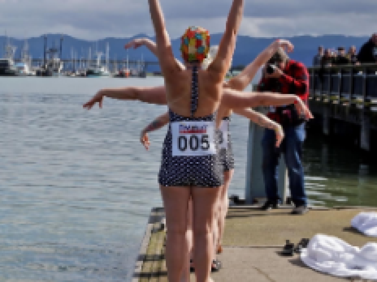 A group of swimmers in swimsuits lined up by the water, raising their arms in synchronized fashion.