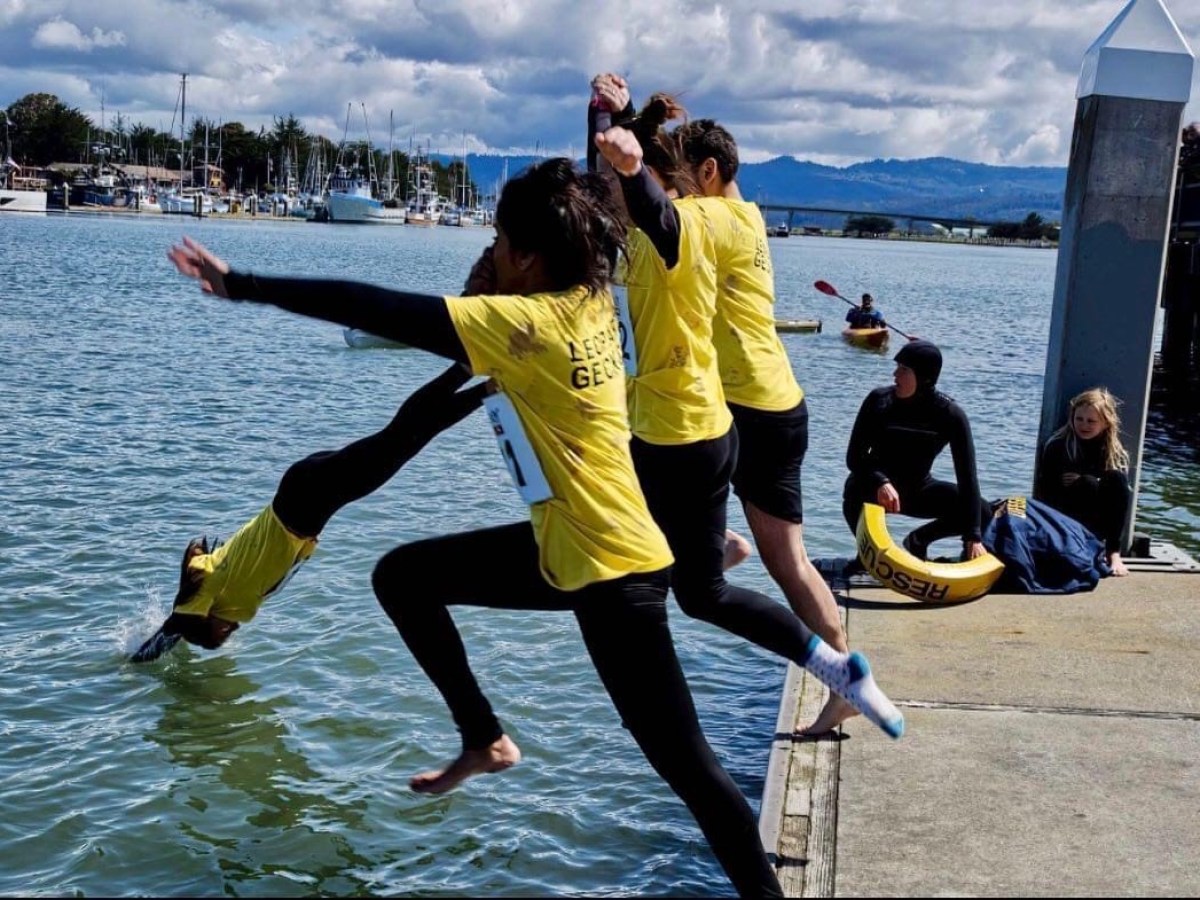 Three people in yellow shirts jump into a marina while another sits watching.